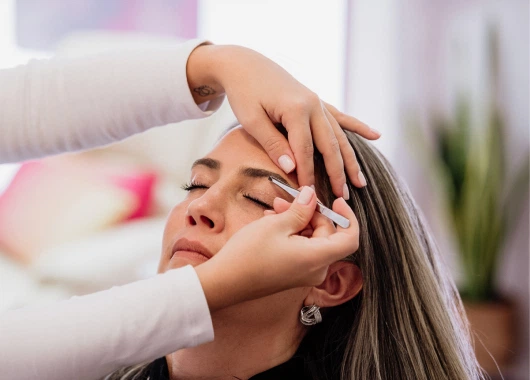 Primer plano de una profesional trabajando en la definición de cejas de una cliente, utilizando pinzas con precisión en un ambiente iluminado y relajado.