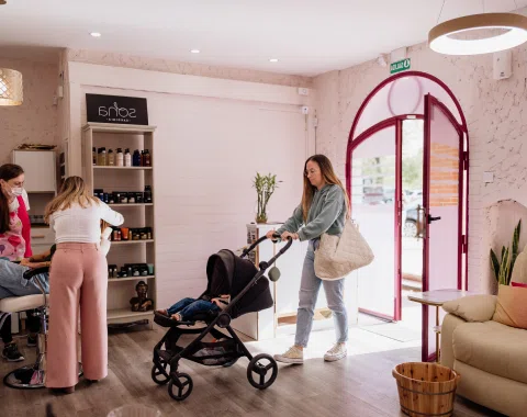Madre entrando al espacio con un carrito de bebé, transmitiendo un ambiente cálido y accesible para familias. Al fondo, estanterías con productos de belleza y cuidado personal.