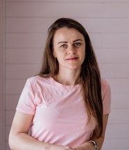 Mujer de cabello castaño largo, con una camiseta rosa claro, sonriendo de manera relajada en un ambiente minimalista.