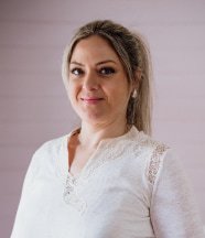 Mujer de cabello rubio sonriente, vestida con blusa blanca de encaje, posando en un entorno luminoso y acogedor.