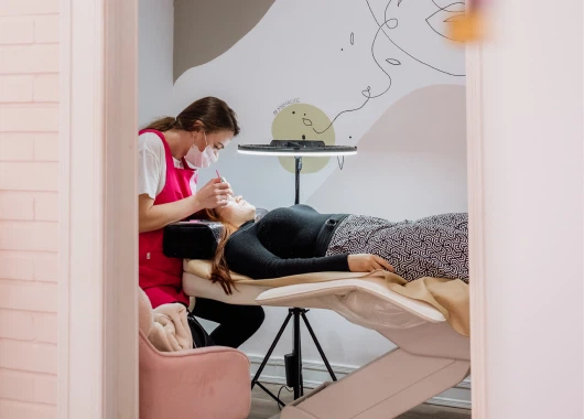 Profesional aplicando extensiones de pestañas a una cliente recostada en una camilla, en un ambiente tranquilo y decorado con elementos artísticos.