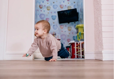 Bebé gateando en el suelo de un espacio decorado con un fondo de globos aerostáticos coloridos y juguetes visibles, transmitiendo un ambiente amigable y pensado para niños.