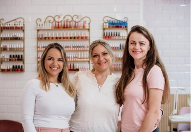 Tres profesionales sonrientes del equipo de Mamá Chic Estética, posando frente a una pared decorada con esmaltes de uñas en un ambiente cálido y acogedor del salón de belleza.