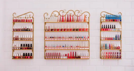 Estantes decorativos llenos de esmaltes de uñas en diversos colores y tonos, organizados en un fondo de azulejos blancos, destacando opciones para todos los gustos.